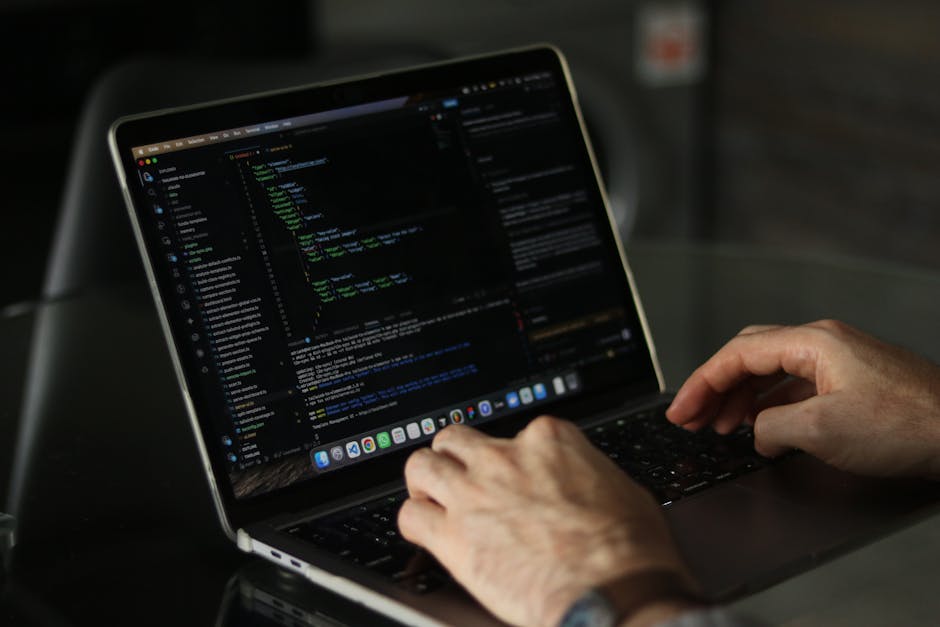 A person typing on a laptop displaying a dark-themed coding interface, with visible lines of green, blue, and white text. The individualâ€™s hands are positioned over the keyboard, and the laptop is p