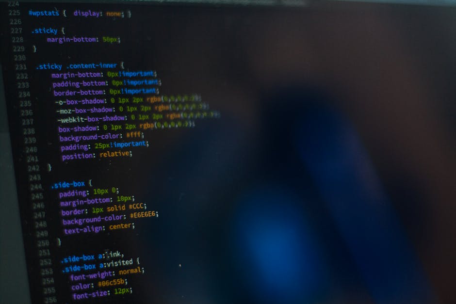 A person typing on a laptop displaying a dark-themed coding interface, with visible lines of green, blue, and white text. The individualâ€™s hands are positioned over the keyboard, and the laptop is p