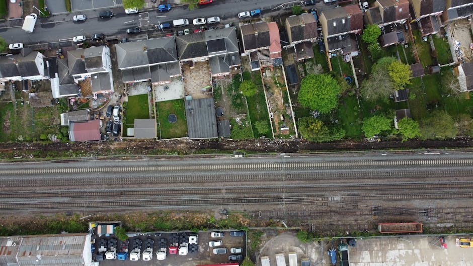 An aerial view capturing a row of small urban backyards separated by fences, with varying features such as lush green lawns, flower beds, and paved patios. Several gardens include trimmed hedges and m