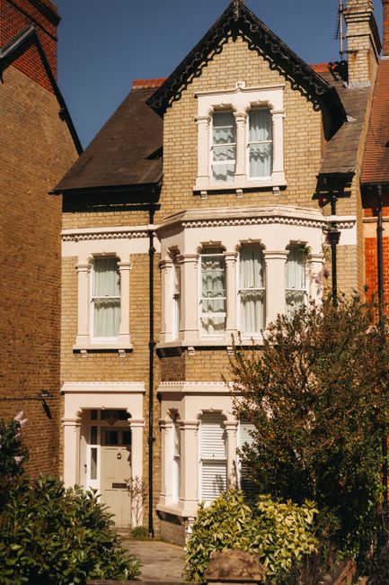 A three-story Victorian-style house with a yellow brick facade and ornate white window frames, situated on a quiet street. The front garden features a variety of green shrubs and small trees, with som